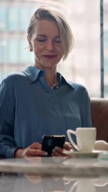 Profesyonel iş kadını kafede otururken telefon kullanıyor. Başarılı girişimci kadın akıllı telefon uygulamasında sosyal medyayı kullanarak kahve içiyor.