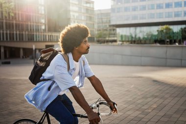 Young African American hipster man with bag riding bicycle on city street going to work 