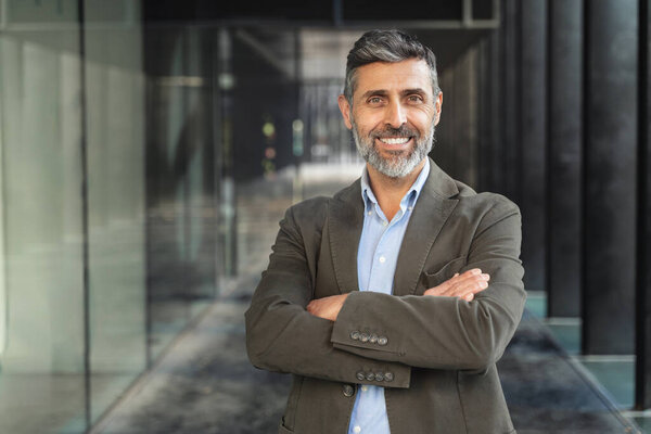 Portrait of mature Spanish businessman standing in the office smiling with crossed arms and positive emotion