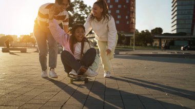 Üç neşeli çoklu etnik arkadaş şehirde gün batımında kaykayla kız iterken eğleniyorlar.