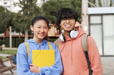 Sırt çantalı ve günlük giysileri olan iki Asyalı öğrenci üniversite kampüsünün önünde gülümsüyor.