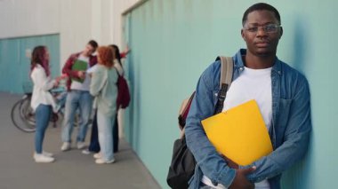 Genç Afrikalı Amerikalı erkek üniversite öğrencisi sınıf arkadaşlarıyla konuşuyor sırt çantasıyla kameraya bakıyor ve gülümsüyor. 