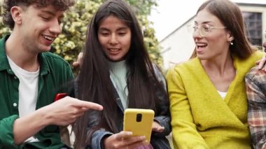 Bir grup genç Gen Z arkadaş cep telefonu uygulamasını kullanarak birlikte oturup komik sosyal medya içeriğini paylaşıyorlar.