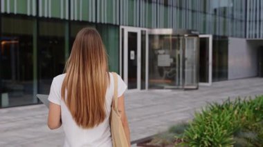 Kitapları olan sarışın kız üniversite binasına doğru yürüyor. Mutlu gülümseyen kadın ileri doğru kameraya bakıyor. 