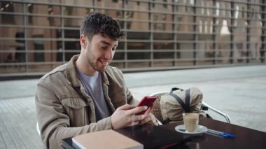 Cep telefonu uygulaması kullanan ve açık kafede gülümseyen genç bir erkek öğrencinin portresi.