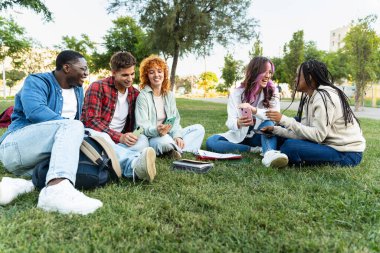 Kampüsteki çimenlerde oturan arkadaşlar, cep telefonlarını sosyal medyada paylaşarak gülüyorlar.
