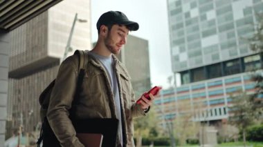 Genç adam cep telefonu kullanırken yürüyor. Şehirde gülümseyen mutlu üniversite öğrencisi