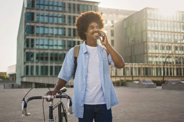 Modern bir şehirde cep telefonuyla konuşurken bisiklet süren gülümseyen Afrikalı Amerikalı adam.