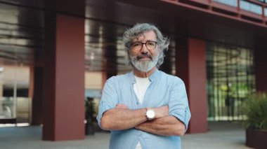 Şehirdeki modern bir binanın önünde kollarını kavuşturmuş yaşlı bir adamın portresi.