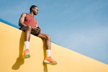 Afrikalı Amerikalı adam modern sarı duvarda oturuyor, spor, antrenman ve koşudan sonra dinleniyor.