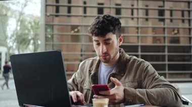 Genç adam açık kafede otururken dizüstü bilgisayar ve cep telefonu kullanıyor.