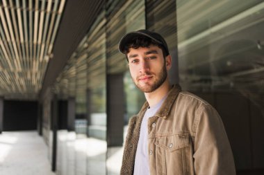 Modern üniversite kampüs koridorunda günlük giysiler ve beyzbol şapkası takan genç bir öğrenci.