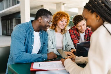 Üniversite öğrencileri bir masada oturup birlikte ders çalışıyor ve dizüstü bilgisayarda ve cep telefonunda yazı yazıyorlar.