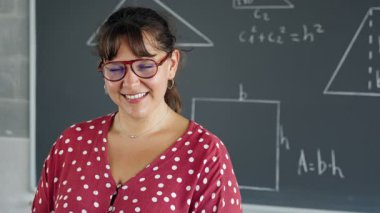 İlkokul sınıfında kameraya gülümseyen gözlüklü genç bir matematik öğretmeni portresi.