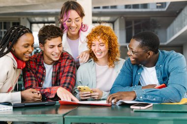 Evde çalışan çeşitli genç öğrenciler cep telefonu ve not defterleriyle birlikte üniversite kütüphanesinde oturuyorlar.