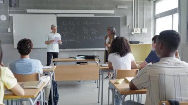 Erkek öğrenci, lise sınıf arkadaşlarına ve öğretmenlerine ödev sunumu okuyor.