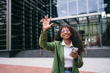 Afrika kökenli Amerikalı girişimci iş kadını cep telefonu tutuyor modern iş şehirlerinde kameralara bakıyor ve olumlu bir ifadeyle selamlıyor.