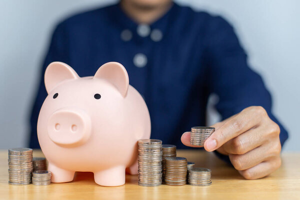 Man hand putting money coins to row of coins with piggy bank on wooden table. Concept for invest and saving success in the future goal