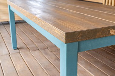 Yemek masası çam ağacından yapılmıştır ve mavi bacakları yakından görülür. Ahşap döşemeli büyük bir masa ve yakınlardaki çimenlik alanı görüyor.