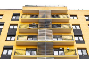 Modern apartman binaları, sarı cepheli, büyük pencereli, metal parmaklıklı balkonlar, bulutlu gökyüzüne karşı duran, alçak açılı manzaralı.