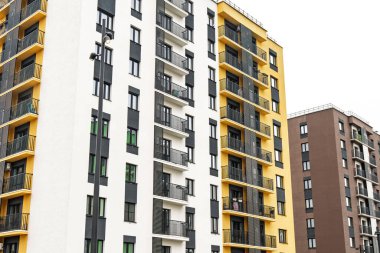 Modern apartmanlar, sarı beyaz ve kahverengi cepheli binalar, pencereler ve metal parmaklıklı balkonlar, bulutlu bir gökyüzüne karşı, kentsel yaşam