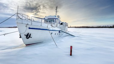 White ship moored in a frozen, snow-covered river, surrounded by a serene winter landscape under a cloudy sky at sunset clipart