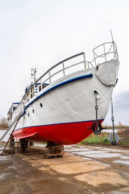 Beyaz ve kırmızı gemi nehir kenarındaki kuru rıhtımda bakım için kaldırıldı. Arka planda bulutlu gökyüzü ve sonbahar ağaçları var. Gemi onarımı kavramı