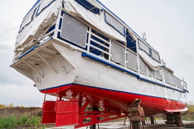Ahşap destekli büyük teknenin kıçı, üst güverte ve metal korkulukları kapalı, bulutlu gökyüzüne karşı alçak açı manzaralı kuru rıhtımda onarım için destekleniyor.