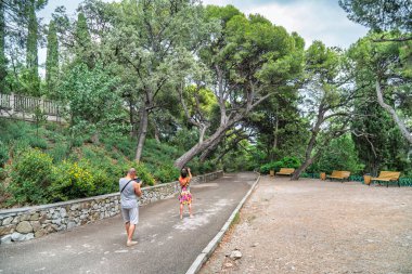 Kadın ve erkek, bulutlu bir günde ağaçların, duvarların, bankların ve yemyeşil çimlerin olduğu manzaralı bir parkta fotoğraf çekiyorlar.