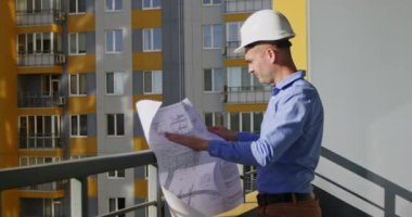 Mimar-tasarımcı, konut kompleksinin yapısını inceler ve çizimleri inceler. Yetişkin adam kağıt üzerindeki grafikleri ve gerçek apartman bloklarını karşılaştırmaya çalışıyor