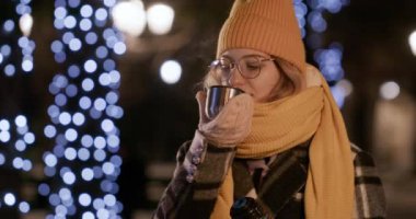 Şapkalı ve eşarplı kadın termostan sıcak çay içmeye çalışıyor. Gözlüklü kadın caddede bulanık ışıklara karşı duruyor.
