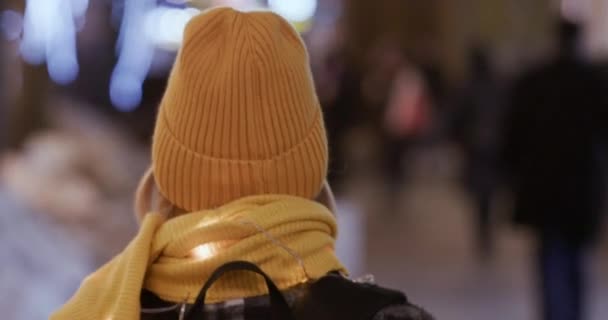 オレンジ色のニットの手作りの帽子とスカーフの女性は ぼやけた背景に照らされた通りを歩く 寒い冬の夜を楽しむ女性の裏側クローズアップ — ストック動画