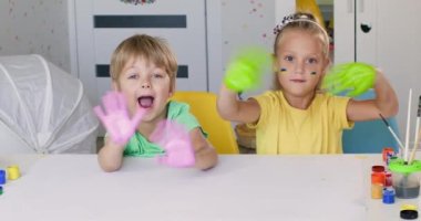 Anaokulu öğrencisi kız kardeş ve küçük kardeş el sallıyor ve odada otururken birlikte gülümsüyorlar. Kardeşler hafta sonları evde vakit geçirmeyi severler.