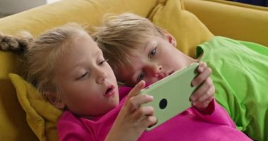 Küçük çocuk akıllı telefondan ablasının oyun oynamasını izlemekten hoşlanıyor. Anaokulu kızı endişeli bir ifadeyle telefona bakar ve oyunu yenmeye çalışır.