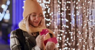 Gözlüklü meraklı kadın pembe hediye kutusunu açıyor ve şaşırmış görünüyor. Bayan Noel ışıklarının karşısında dikildiği için çok mutlu.