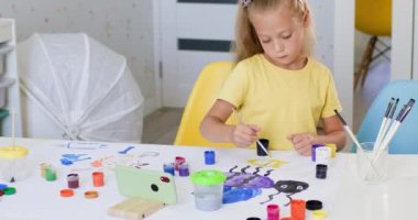 Preschooler girl enjoys painting pictures with bright gouache. Child with excited and amused expression prepares big painting on table