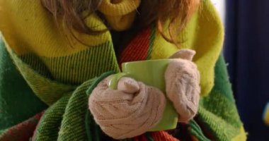 Lady in winter mittens warms up from hot mug with beverage and wants to drink. Woman covered with blanket tries different ways to warm in cold apartment
