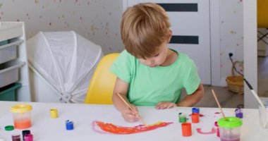 Excited toddler boy puts effort into painting nice picture at home. Kid wearing green t-shirt paints rainbow arc with watercolor paints on paper