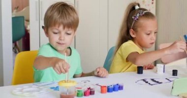 Excited siblings paint pictures together with gouache on paper sheet in apartment. Sister and brother sit at table and enjoy painting picture