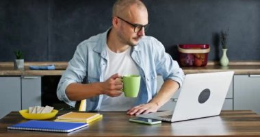 Adult man freelancer drinking hot beverage from mug works on laptop and types information about project. Businessman enjoys working from home office