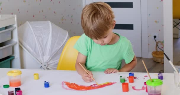 興奮した幼児の男の子は家で素敵な絵を描くことに努力を置きます 緑のTシャツを着た子供は 紙の上に水彩画で虹の弧を描きます — ストック動画