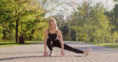 Young woman wearing black sportsuit stretches body for running in autumn park. Sportswoman does exercises for legs squatting on empty pavement