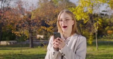 Şok olmuş ve heyecanlı ifadeli sarışın kadın akıllı telefonun çığlıklarına bakıyor. Sarı saçlı bayan sonbahar parkında mutlu haberlerden hoşlanıyor.