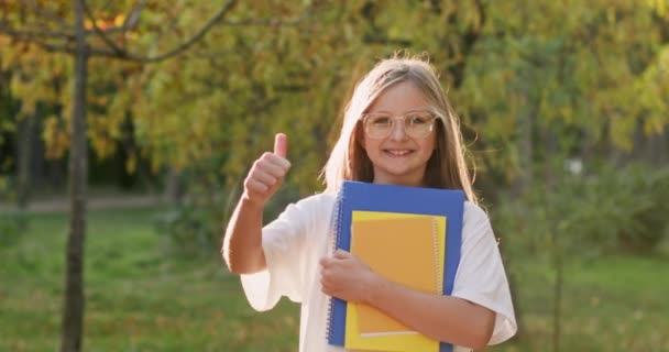 拿着抄本的少女在秋天的树上露出了微笑的大拇指 年轻的金发姑娘对开学很兴奋 — 图库视频影像