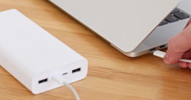 Adam, ahşap masadaki kabloyla güç bankasını bilgisayardan dizüstü bilgisayara bağlıyor. Bilgisayarı şarj etmek için taşınabilir cihaz. Elektrik ekipmanları