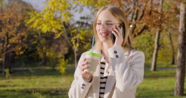 Genç bir kadın, kağıt bardaktan kahve içer ve arkadaşıyla akıllı telefondan konuşur. Sarı saçlı bayan sonbahar parkında yürüme duygularını paylaşıyor.