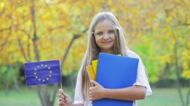 Gülümseyen yüzlü sarışın genç elinde AB bayrağı ve defterleri tutuyor. Uzun saçlı kız öğrenci yurtdışında okumaya başlıyor. Pozitif duygular gösteriyor.