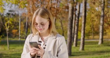 Gülümseyen ve mutlu yüzlü genç bir kadın akıllı telefonlara gülerek bakıyor. Gözlüklü sarışın kadın utangaç ve sıcak sonbahar havasından hoşlanıyor.