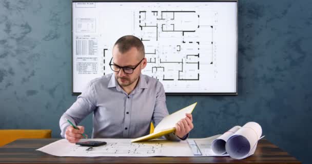 エンジニアは計算機を使用して計算を行い 図面に結果を書きます 建築家は大きな図面に対してテーブルに座って黄色の紙のメモ帳にメモを取ります — ストック動画