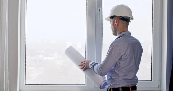 Kasketli ve ofis elbiseli yetişkin mühendis el çizimi ile odayı karşılaştırır. İşçi pencerenin yanında durup etrafı inceliyor.